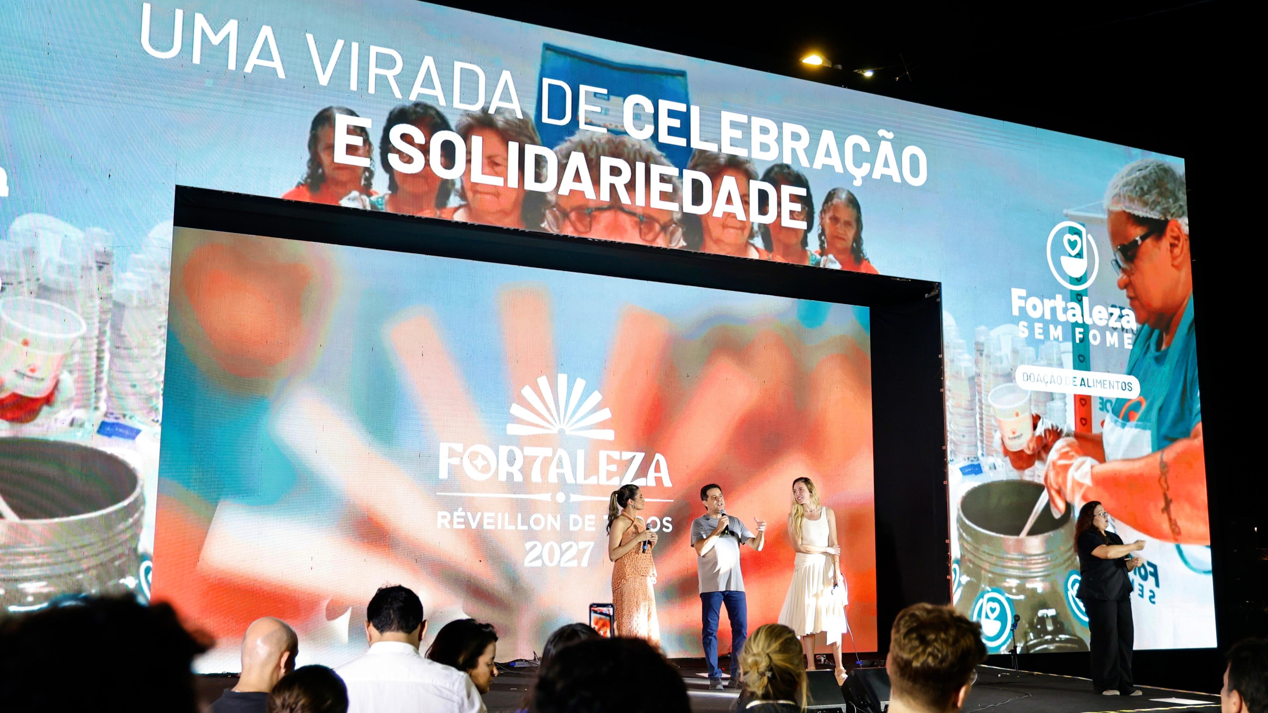 A foto mostra um grande palco com transmissão em led sobre o qual estão o prefeito Evandro Leitão, a secretária do Turismo Denise Carrá e a vice-governadora Jade Romero 
